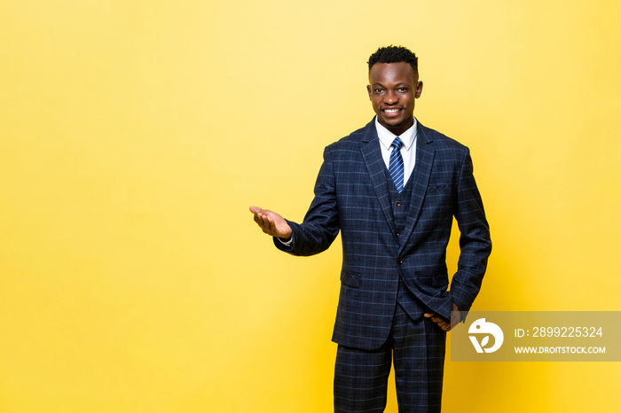 Positive African man in formal business suit smiling and opening hand doing welcome gesture in yellow color studio isolated background