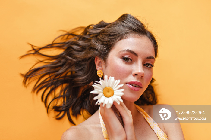 Close up portrait of elegant young caucasian girl on yellow background. Her brown wavy hair is beautifully laid out to sides. Small chamomile near her face.