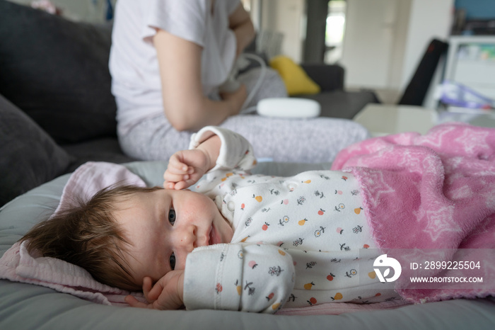 Small caucasian baby infant sleeping on the couch sofa bed at home while her mother in the background is using breast pump to provide milk for feeding