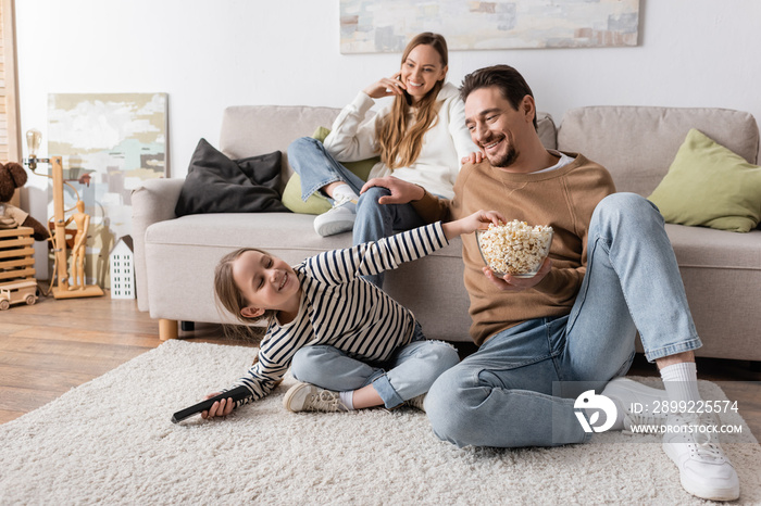 happy kid with remote controller reaching popcorn near father and mother on blurred background.