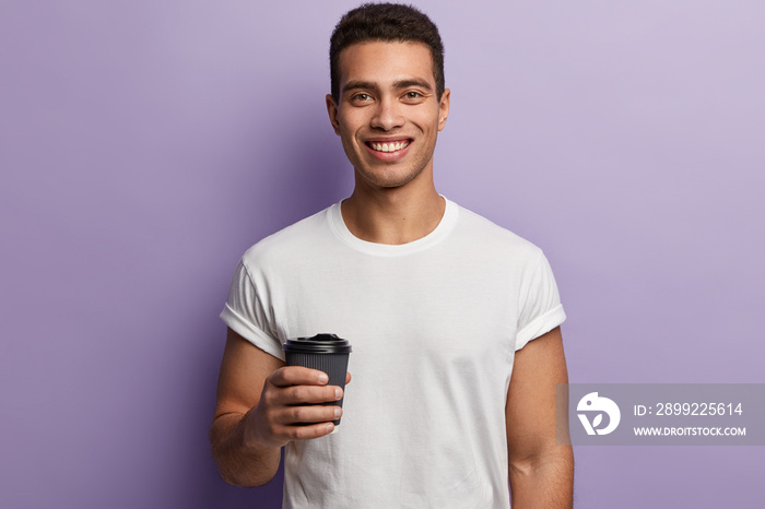 Young hipster male with toothy smile, enjoys free time, wears casual white t shirt, holds takeaway coffee, isolated over purple background, drinks hor beverage after lunch. People and leisure