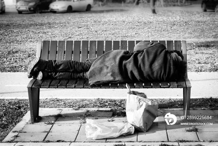 Homeless refugee man in black coat sleeps on the bench on the street black and white