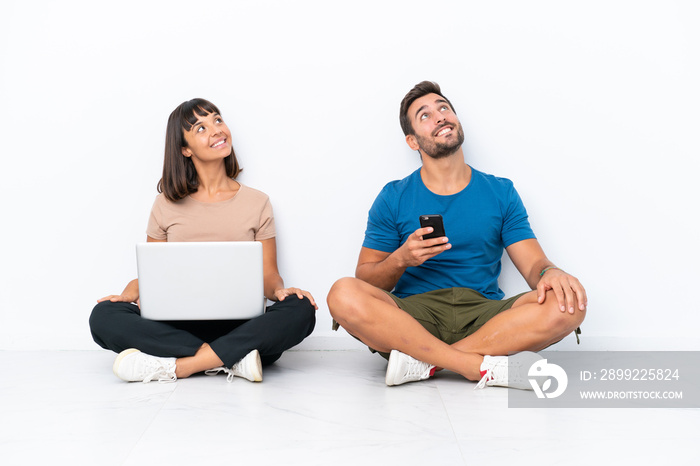 Young couple sitting on the floor holding pc and mobile phone isolated on white background looking up while smiling