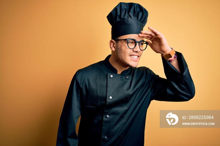 Young brazilian chef man wearing cooker uniform and hat over isolated yellow background very happy and smiling looking far away with hand over head. Searching concept.