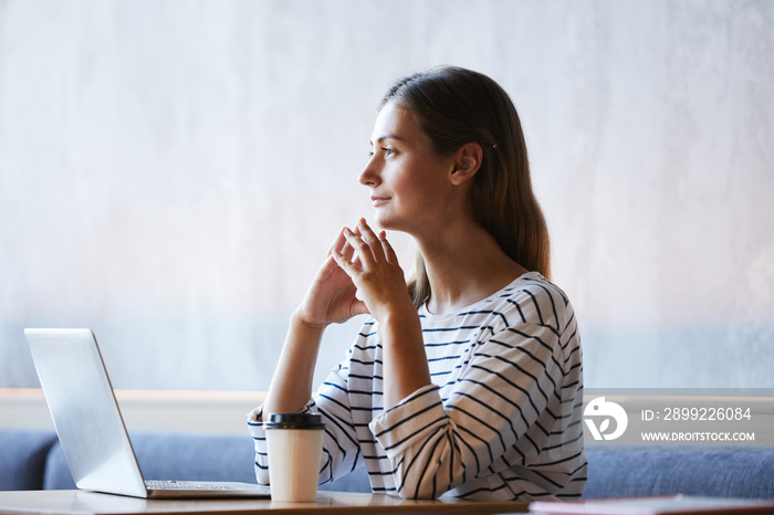 Introspective purposeful young lady sitting at table in modern cafe and thinking of new internet project