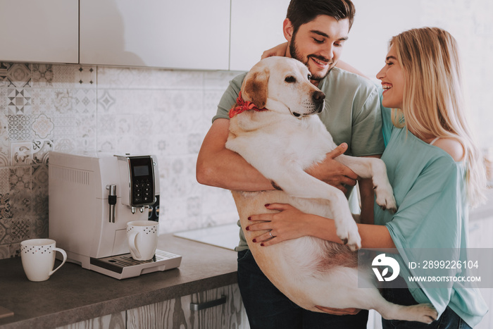 Waist up portrait of beautiful happy woman embracing her handsome bearded boyfriend while he holding large dog. They are standing in the kitchen near the coffee machine