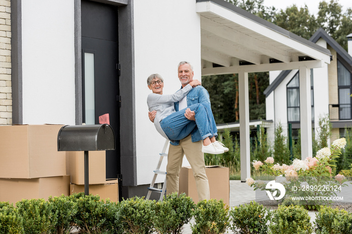 mature man holding smiling woman in glasses near new house