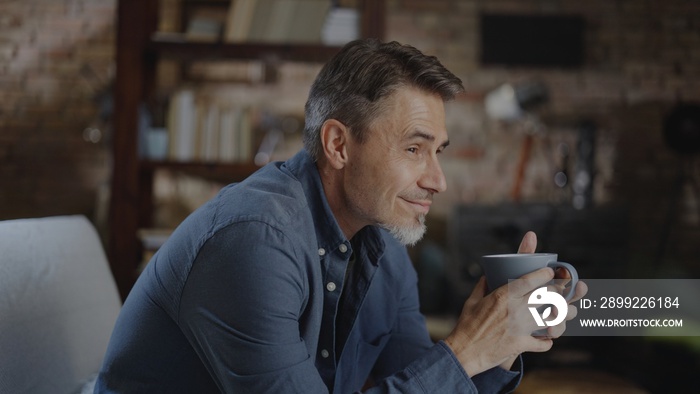 Confident older white man at home, sitting on couch in dark living room, daydreaming, drinking coffee or tea. Portrait of happy, mature age, middle age, mid adult caucasian man in 50s, smiling.