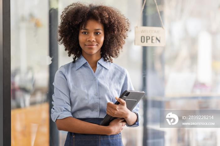 Successful african woman in apron standing coffee shop door. Happy small business owner holding tablet and working. Smiling portrait of SME entrepreneur seller business standing with copy space.