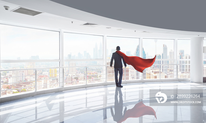 Businessman in a suit and hero cape and looking at the city, stands in empty office. Victory concept, morning scene .