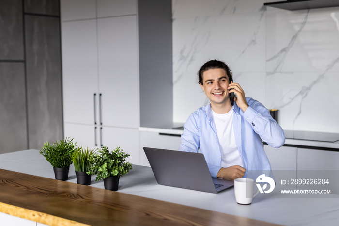 Gig economy concept. Young positive man freelancer working remotely at home, looking at laptop and consulting client via cellphone, sitting at kitchen