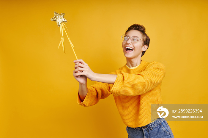 Playful young woman holding magic wand while standing against yellow background