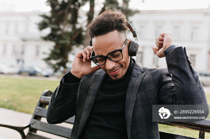 Pleased dark-haired african man wears black clothes chilling outdoor. Photo of relaxed guy in glasses enjoying music in headphones.