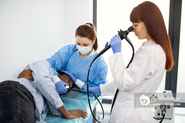 Female concentrated doctor gastroenterologist, operating endoscope during gastroscopic procedure of young afro-american man patient, lying on the couch