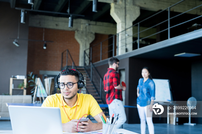 Portrait of  creative young Middle-Eastern man wearing headphones and bright yellow shirt using laptop and listening to music while working in open space office of IT developers team, copy space