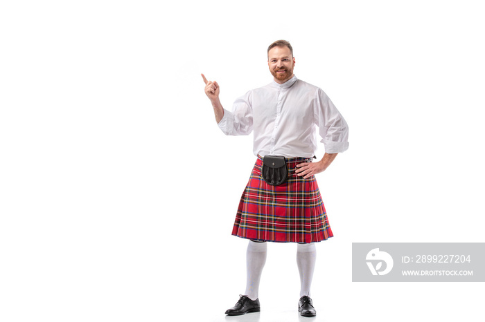 smiling Scottish redhead bearded man in red tartan kilt pointing with finger on white background