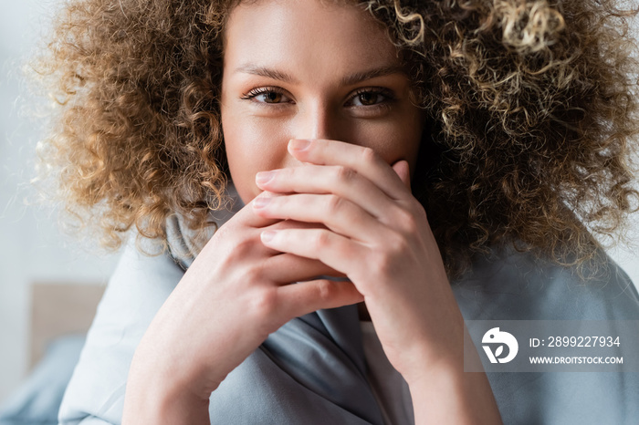 young curly woman with happy eye expression obscuring face with hands and looking at camera.