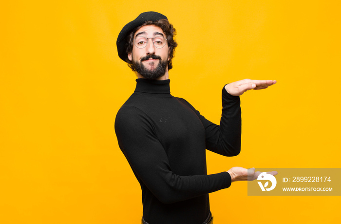 young french artist man smiling, feeling happy, positive and satisfied, holding or showing object or concept on copy space against orange wall