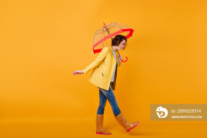 Trendy girl in rubber shoes and yellow jacket looking down while posing with umbrella. Studio shot of curly short-haired woman in jeans walking with parasol.