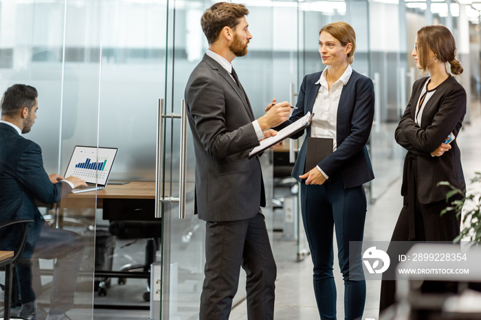 Business people talking in the hallway of the modern office building with employees working behind glass partitions. Work in a large business corporation