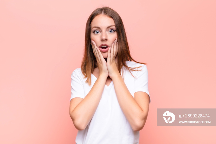 young red head woman feeling shocked and scared, looking terrified with open mouth and hands on cheeks against flat wall