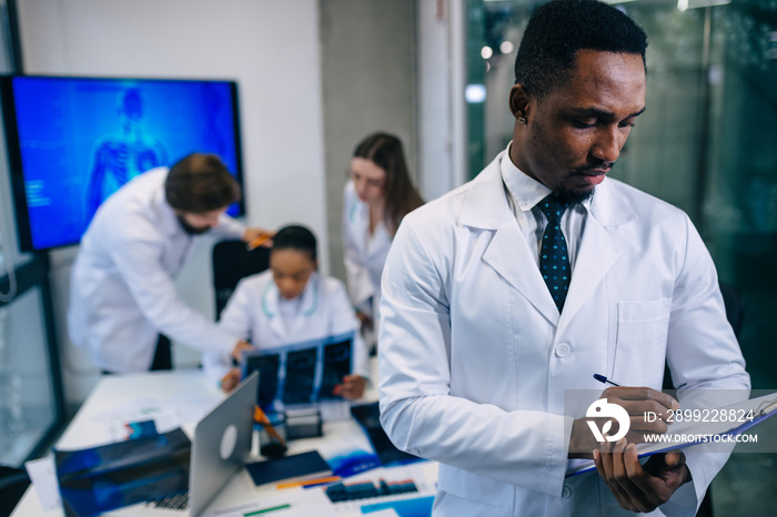 A serious African-American doctor takes notes of the patient’s diagnosis, puts together a plan of treatment, while the colleagues behind them discuss computer diagnostics, MRI, X-rays