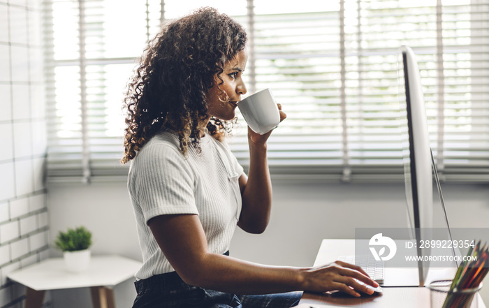 Portrait of smiling happy african american black woman relaxing using technology of desktop computer while sitting on table.Young creative african girl working and drink coffee at home.work from home