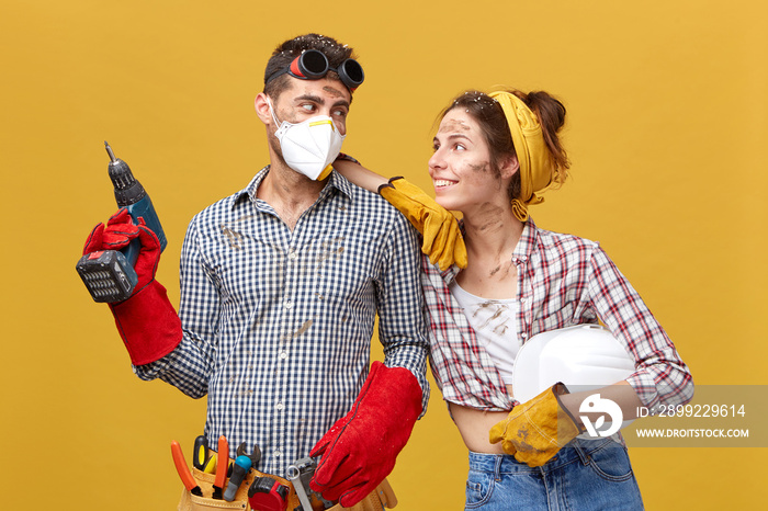 Portrait of foreman and his wife standing against yellow background looking at each other with smile, being busy with builduing. Young male engineer wearing protective mask working with drill