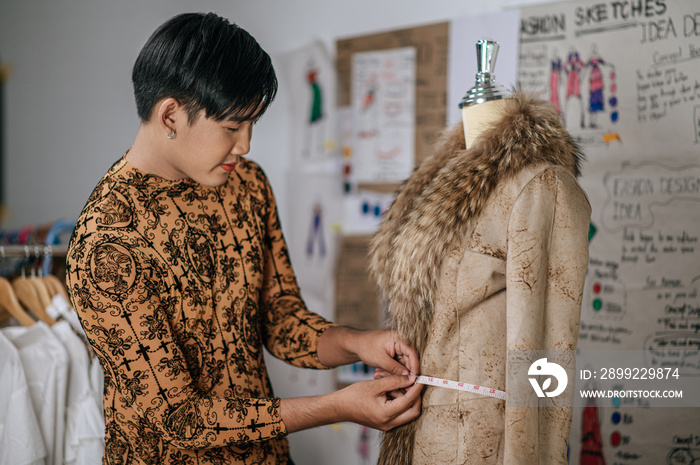 Portrait of Asian young male tailor measuring mannequin’s waist at table in fashion studio. Designer and LGBTQ Concept.