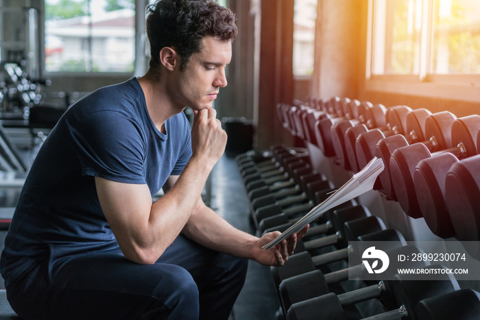 Muscular handsome trainer looking at fitness plan on clipboard for working out in the fitness gym