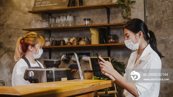 Young Asia female wear face mask self service use mobile phone pay contactless at restaurant. Girl barista talking receive order from customer at cafe. Lifestyle new normal after corona virus concept.