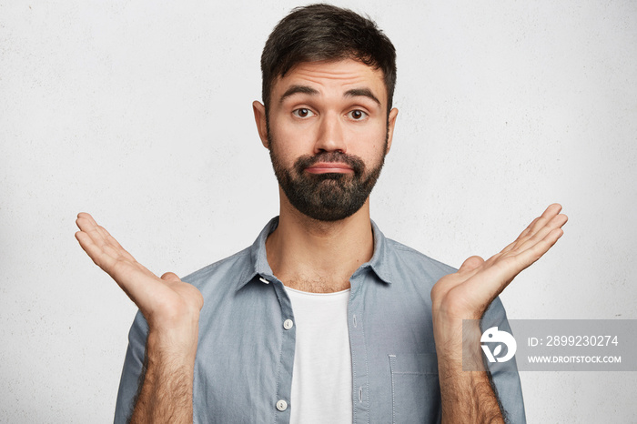 Photo of uncertain bearded male with appealing appearance, dressed casually, shrugs shoulders in doubt, is ignorant of new facts, confused about future plans, isolated over white background.