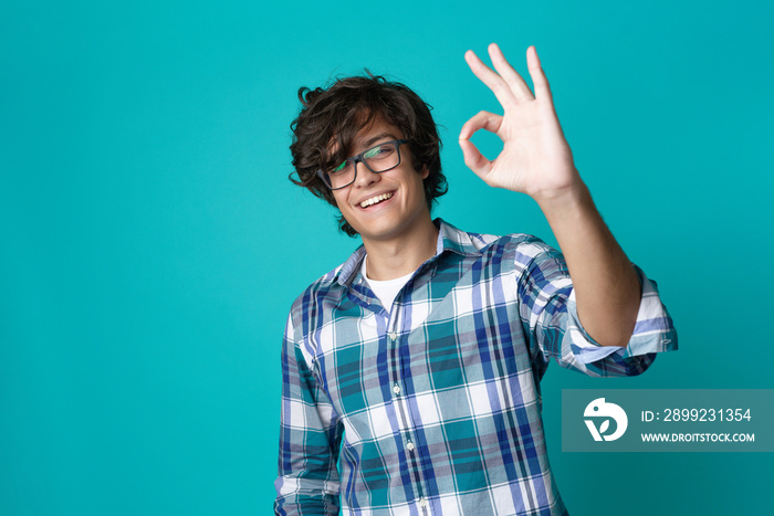 Young man with spectacles making an ok sign, expressing approval and looking cheerful and content; teal studio background