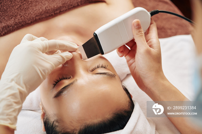 Close-up image of beautician using ultrasound tool for cleaning nose pores of young woman