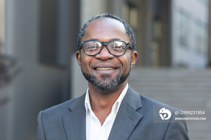 Close-up photo. Portrait of a successful handsome African American man. A businessman, lawyer, boss stands in front of the office center in a suit and glasses, looks at the camera, smiles.