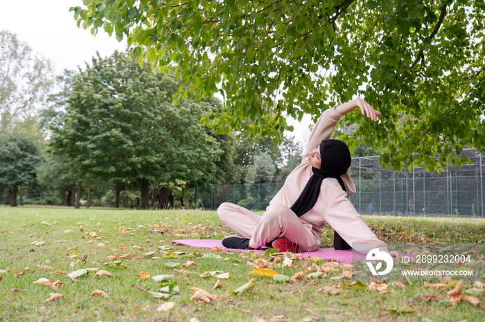 Woman in hijab and pink tracksuit practicing yoga in park