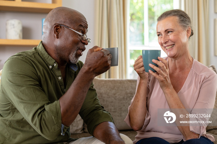 Happy senior multiracial couple having coffee while sitting on sofa at home