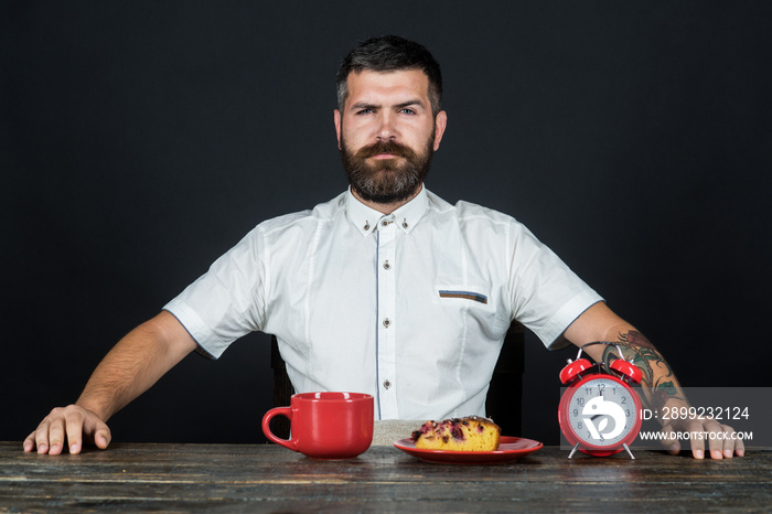 Bearded man sitting at table with cup of morning coffee. Coffee time concept - red alarm clock, cup with coffee and piece of tasty pie on plate. Morning breakfast of coffee and cake on wooden table.