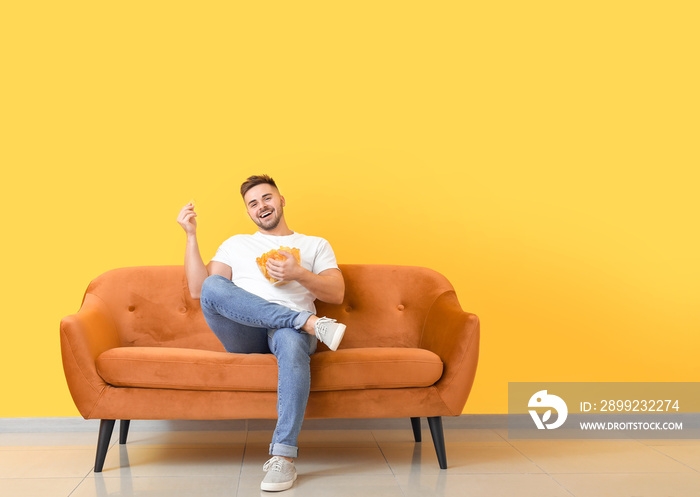 Handsome young man with tasty potato chips sitting on sofa near color wall