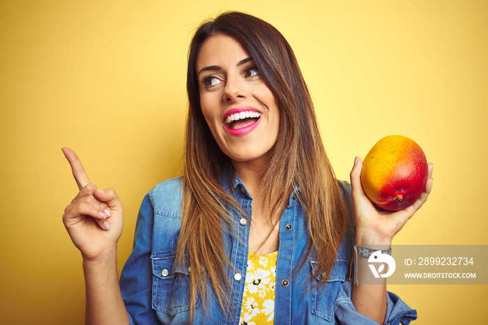 Young beautiful woman eating fresh healthy mango over yellow background very happy pointing with hand and finger to the side