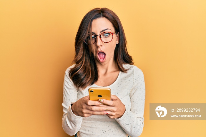 Young brunette woman using smartphone over yellow background afraid and shocked with surprise and amazed expression, fear and excited face.