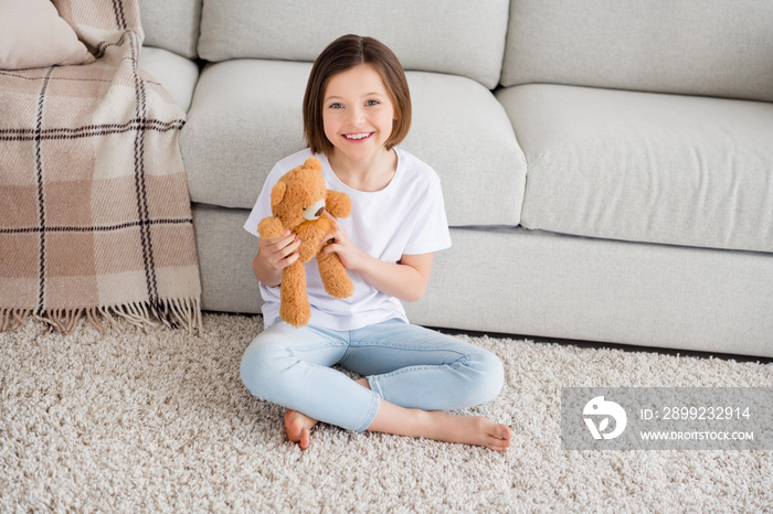 Full length photo of young cheerful small girl happy positive smile sit floor comfort play fluffy bear toy home