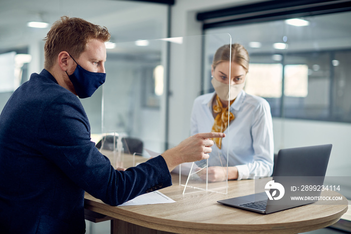 Business colleagues with face masks reading an e-mail on laptop while being separated with sneeze guard in the office.