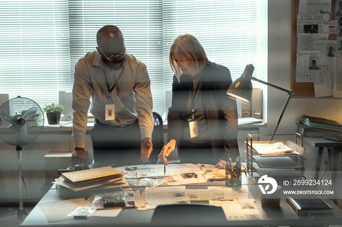 Two confident FBI agents bending over desk with criminal profiles and evidences while pointing at photo of suspect during discussion