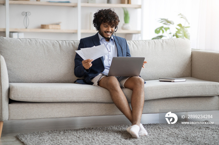 Attractive indian guy sitting on couch, using laptop and headset