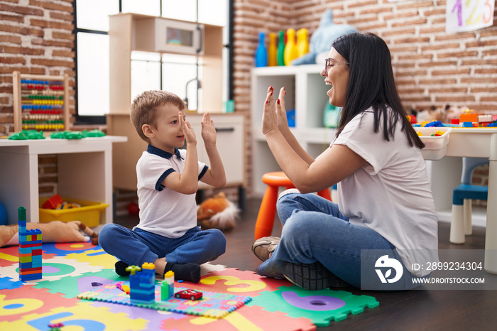 Teacher and toddler sitting on floor high five with hands raised up at kindergarten