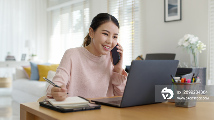 Young happy attractive beautiful asia female staff laugh talk on mobile phone relax comfort sit work on desk table at home. Remotely work on distance job telesale or secretary career for business.