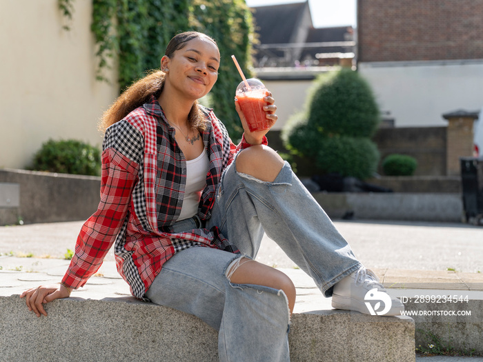 Portrait of young woman with smoothie sitting on stone wall