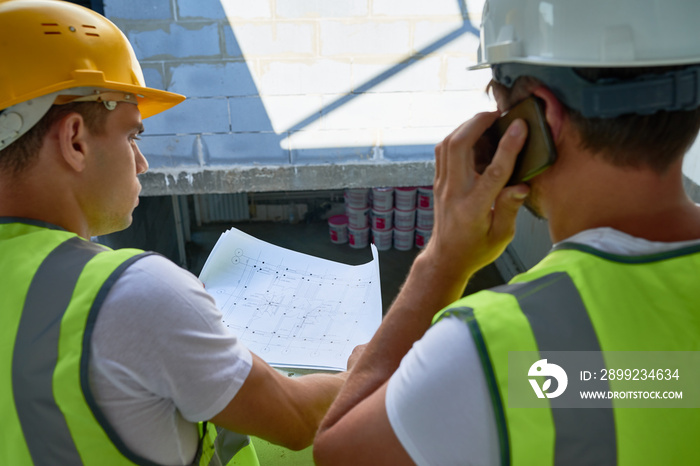 Back view of unrecognizable construction workers wearing reflective jackets and hardhats discussing floor plan outdoors, one of them distracted due to phone call,  unfinished building on background