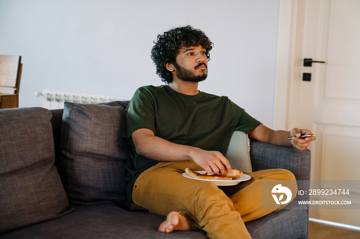 Young indian handsome curly man sitting on sofa switching channels